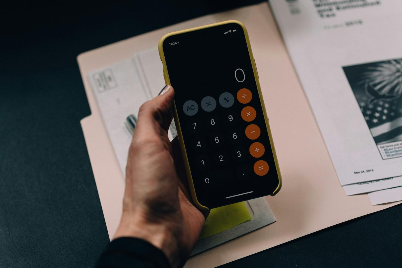 A man holding a smartphone with his calculator app open and some documents on his table, trying to calculate for the costs of hiring a team building speaker.