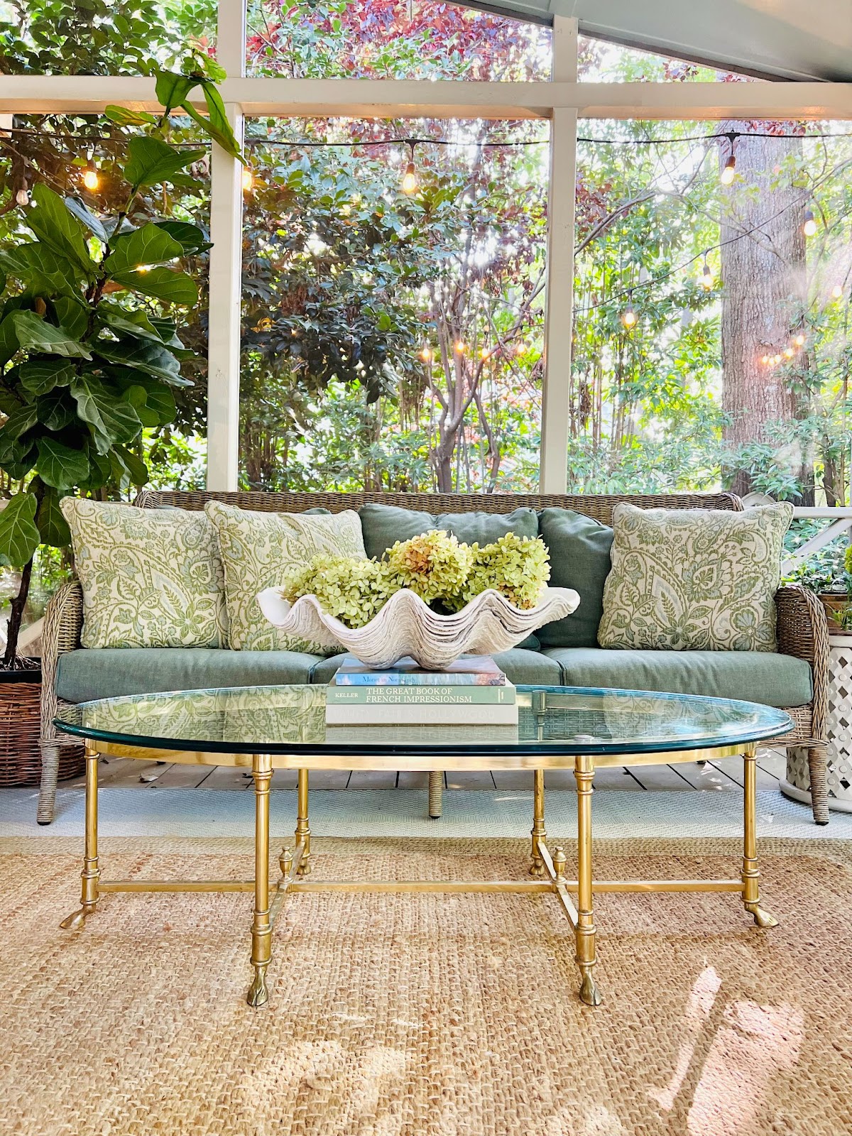 A beautiful sunroom adorned with a cozy rattan sofa, lush greenery, a sleek glass-top table, and upholstery and throw pillows in shades of green.