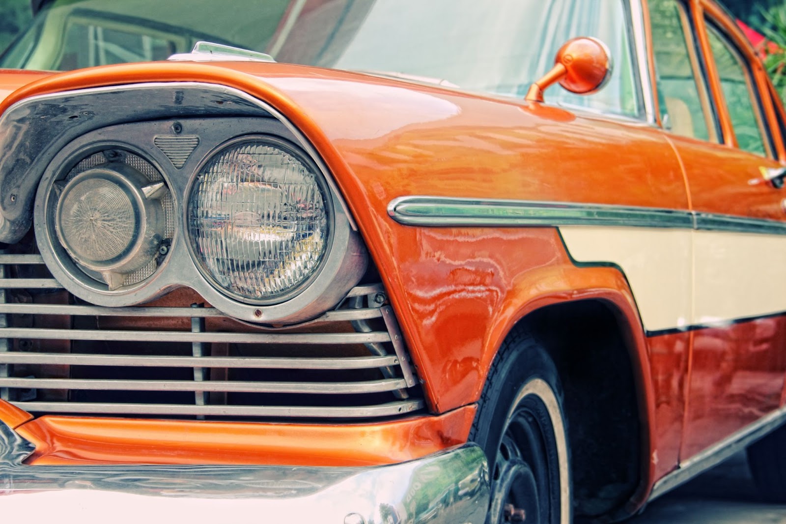 Close up de carro antigo alaranjado, com o seu farol esquerdo em foco e a lateral passando para o fundo da imagem.