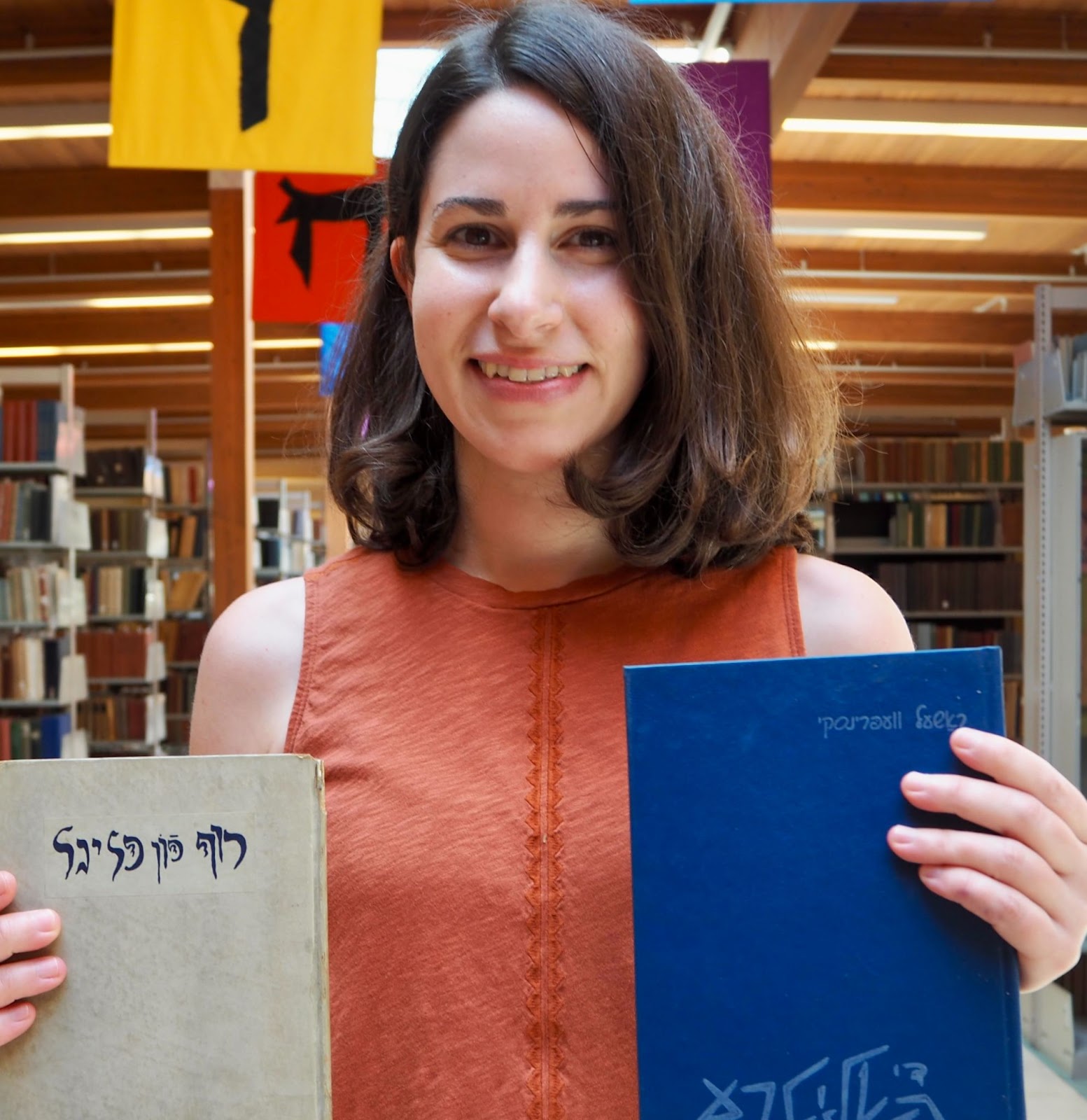Rebecca Weingart, a white woman with brown shoulder-length hair, smiles at the camera while holding two books, one blue and one tan, with Yiddish writing on the covers. She is wearing a sleeveless orange top and is posing in front of a background of bookshelves.