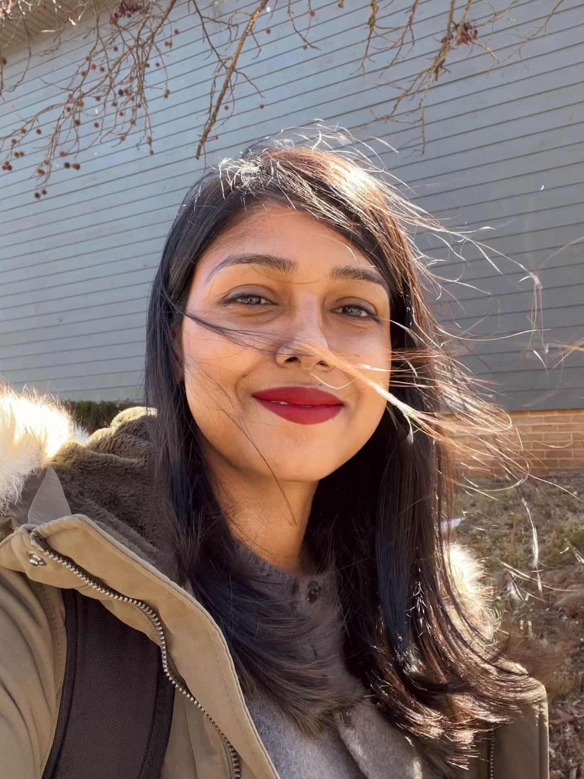 Nidhi, a South Asian woman with black hair, smiles at the camera against the wall of a grey wooden house. She’s wearing an olive winter coat with a fur hood over a grey sweater.