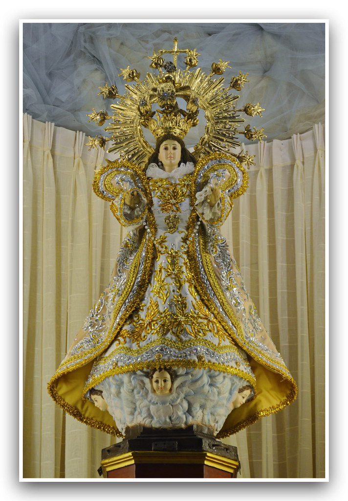 A statue of the Virgin Mary adorned in ornate gold and white robes, with a crown and halo, standing on a cloud with cherubic faces.