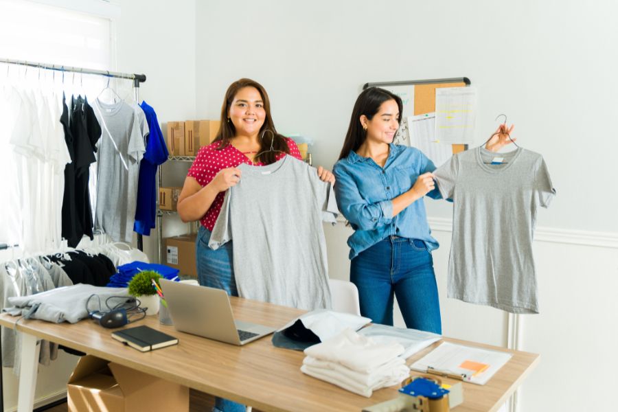 Two women displaying gray t-shirts in a home-based business setup.