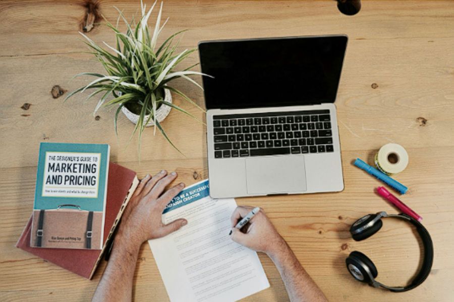 Workspace with laptop, marketing book, and person writing on a document.