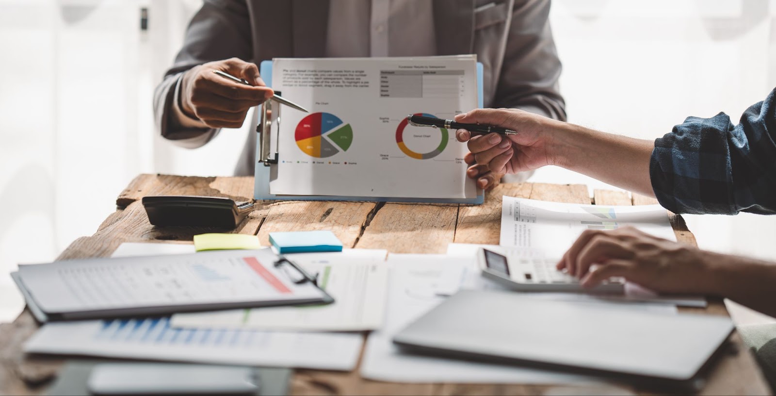 Two colleagues discuss business and analyze price charts, with various documents and charts on the table, illustrating business strategies and budgeting basics.