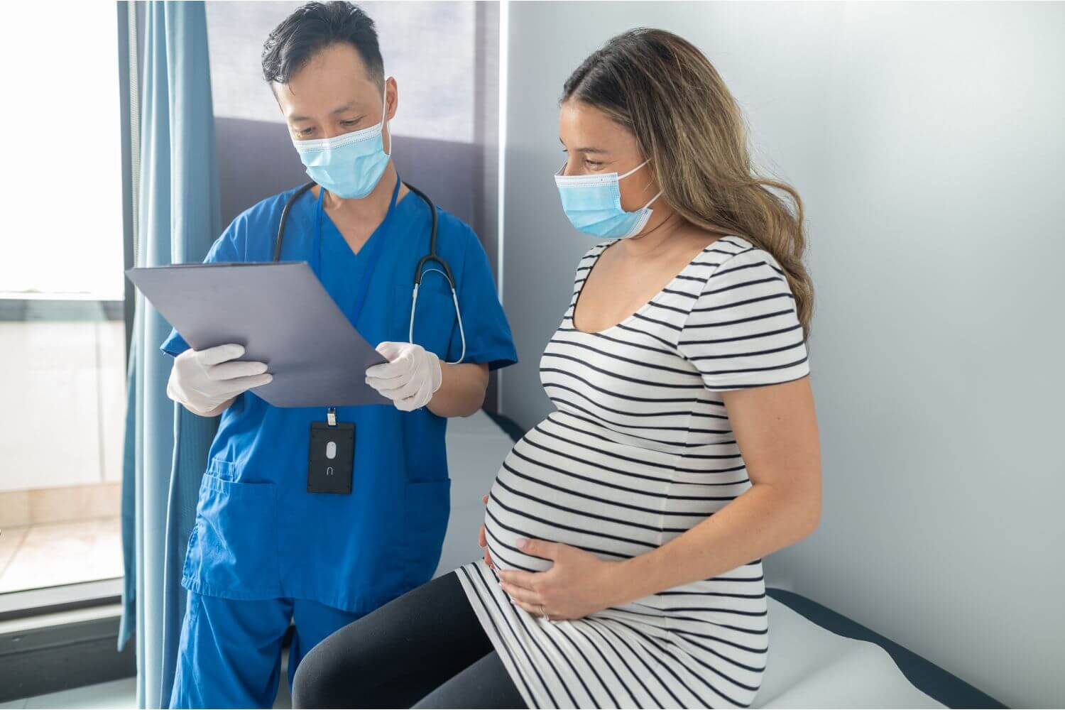 Pregnant woman sitting on an exam table next to her physician holding a clipboard