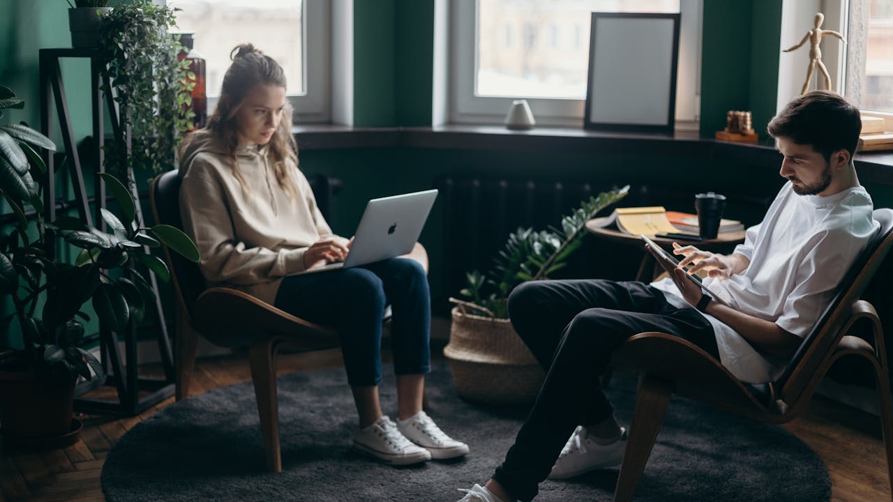 Two people seated in a living room with one working on a laptop, and the other using a tablet.