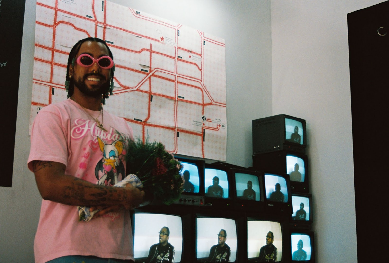 Image: Montalvo smiling alongside a vintage television installation playing a looped video history of Craig Lovinggood, known as drag queen Tisha Taylor. Behind Montalvo and the installation is a map of {B/qKC}’s five public installations displayed in April 2024.