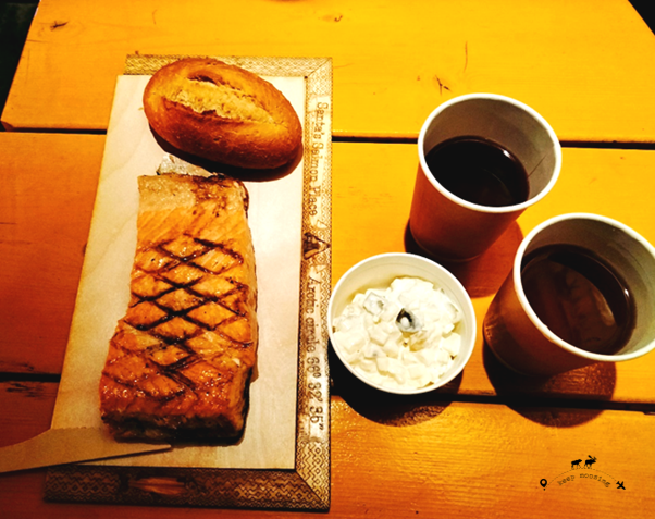 The very delicate salmon of Santa's Salmon together with a piece of bread, an onion sauce and two glasses of blueberry juice. All this placed on a large light wooden table.