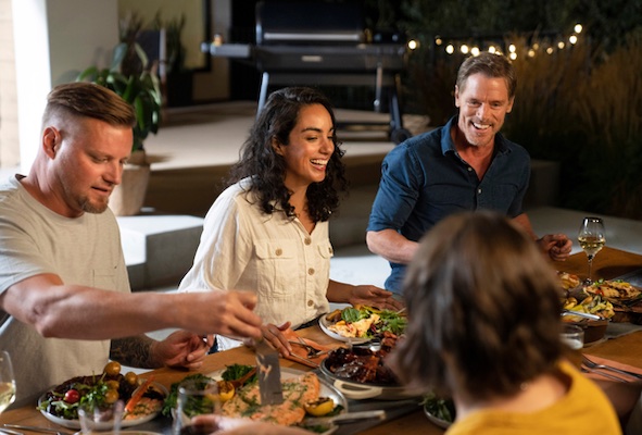 Alt Text: A group of friends at a table eating a meal and sharing laughs outside on a backyard patio with a Traeger pellet grill in the background.