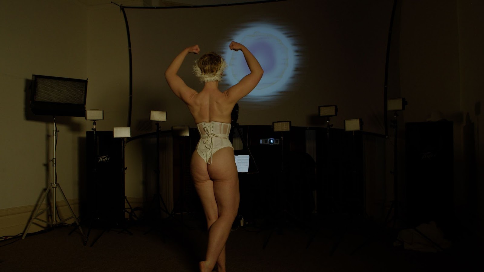 Image: Bun Stout performs “The Swan” at PS1 Close House. A performer wearing a white corset and underwear poses in front of a projector screen with their back turned toward the camera. Their arms are lifted above their head and are flexing, displaying the rippled muscles of their back. A line of unlit lights is arranged in front of the projector screen. A luminous sphere is displayed on the projector screen. Photo courtesy of the artist. 