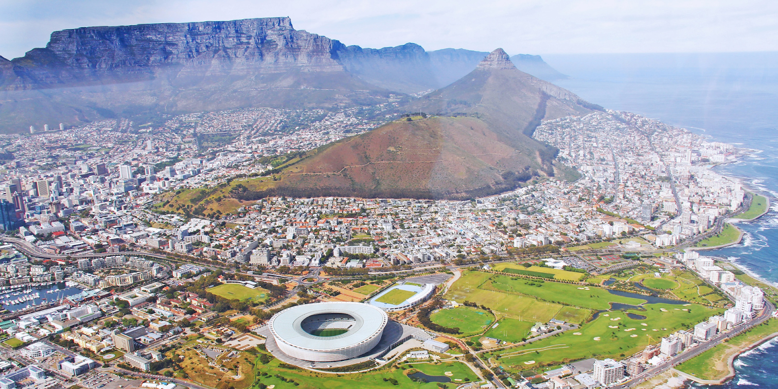 A stunning aerial view of Cape Town, South Africa, featuring the iconic Table Mountain in the background. The city's landscape includes a mix of modern buildings, residential areas, and green spaces. The Cape Town Stadium is prominently visible, with its unique circular design. The coastline and the Atlantic Ocean add to the picturesque scene, showcasing Cape Town's beautiful blend of natural and urban elements.