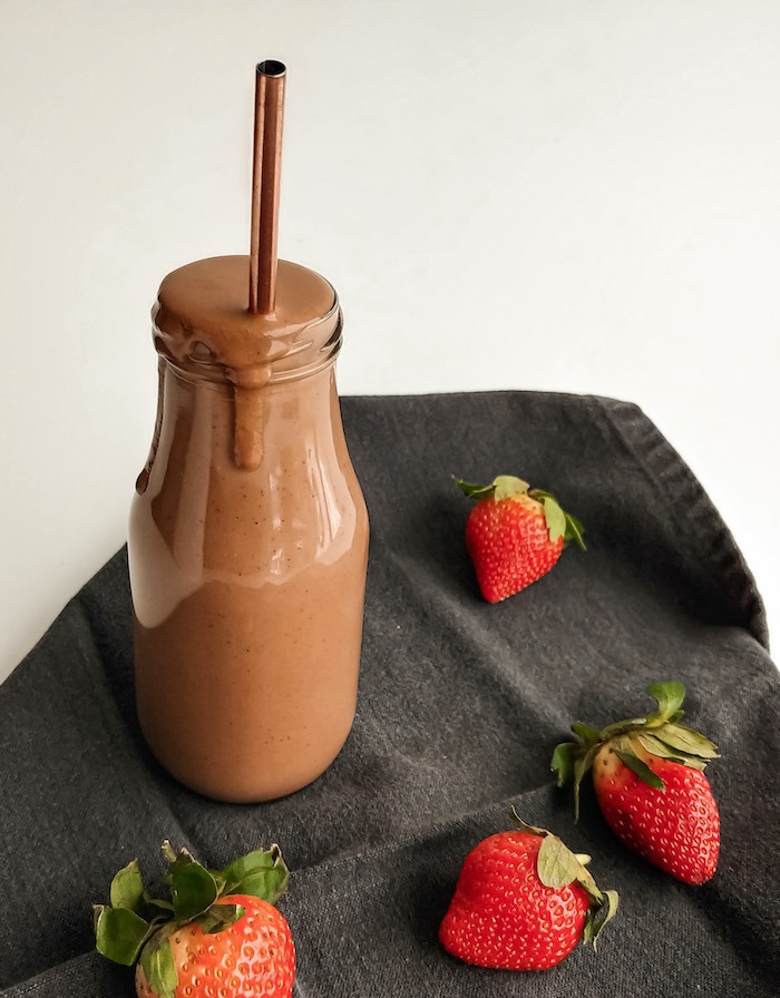 Chocolate and strawberry smoothie in a glass bottle with a metal straw, surrounded by fresh strawberries on a gray cloth.