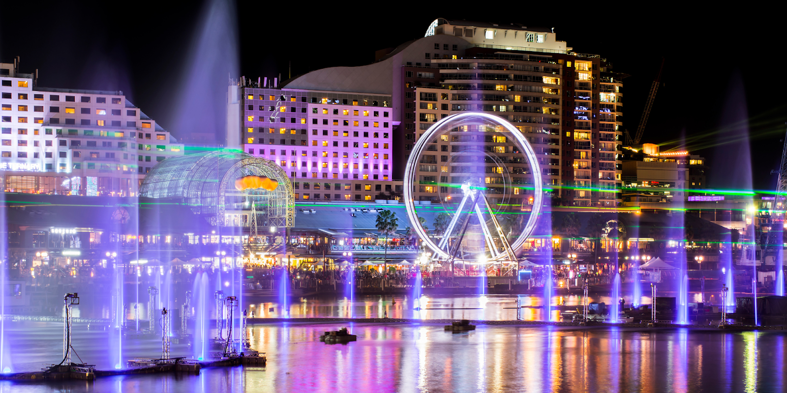 A vibrant night scene at Darling Harbour in Sydney, Australia, featuring a large Ferris wheel illuminated with colorful lights. The buildings surrounding the harbour are lit up, reflecting beautifully on the water. A dynamic light and water show is taking place, adding a festive and lively atmosphere to the waterfront area, with streams of water lit in various colors dancing in sync with the lights.