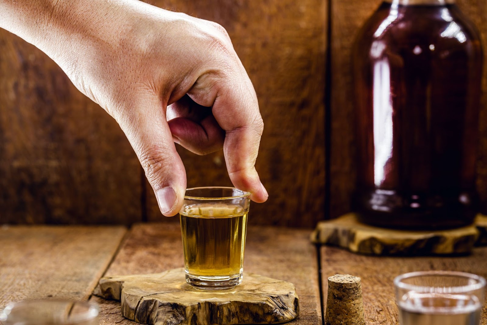 Mão esquerda de uma pessoa prestes a pegar um pequeno copo com uma dose de cachaça.