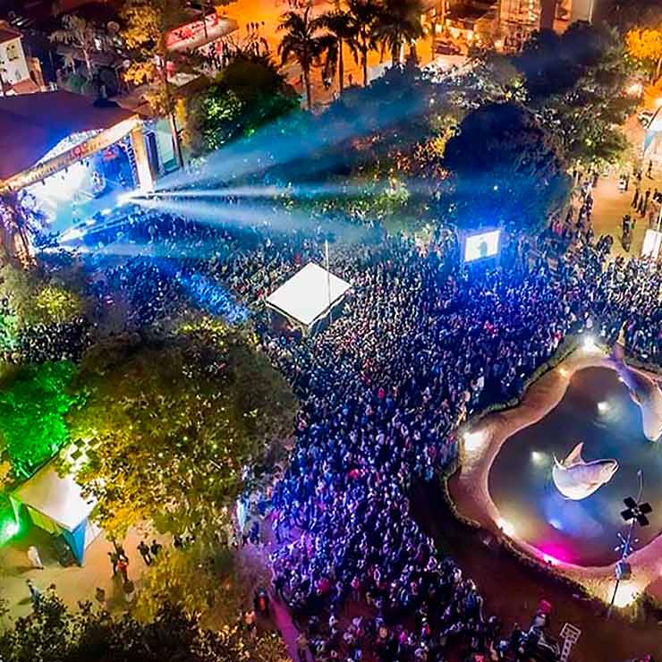 Vista aérea de um grande evento noturno ao ar livre, com multidão, palco iluminado, luzes coloridas e uma fonte decorativa