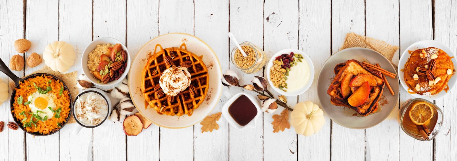 A variety of breakfast foods on a white wooden table, including waffles, pancakes, yogurt with granola, and a skillet dish with an egg. Autumn-themed decorations are scattered around the dishes, sparking thoughts of a Pancakes vs Waffles virtual team debate.