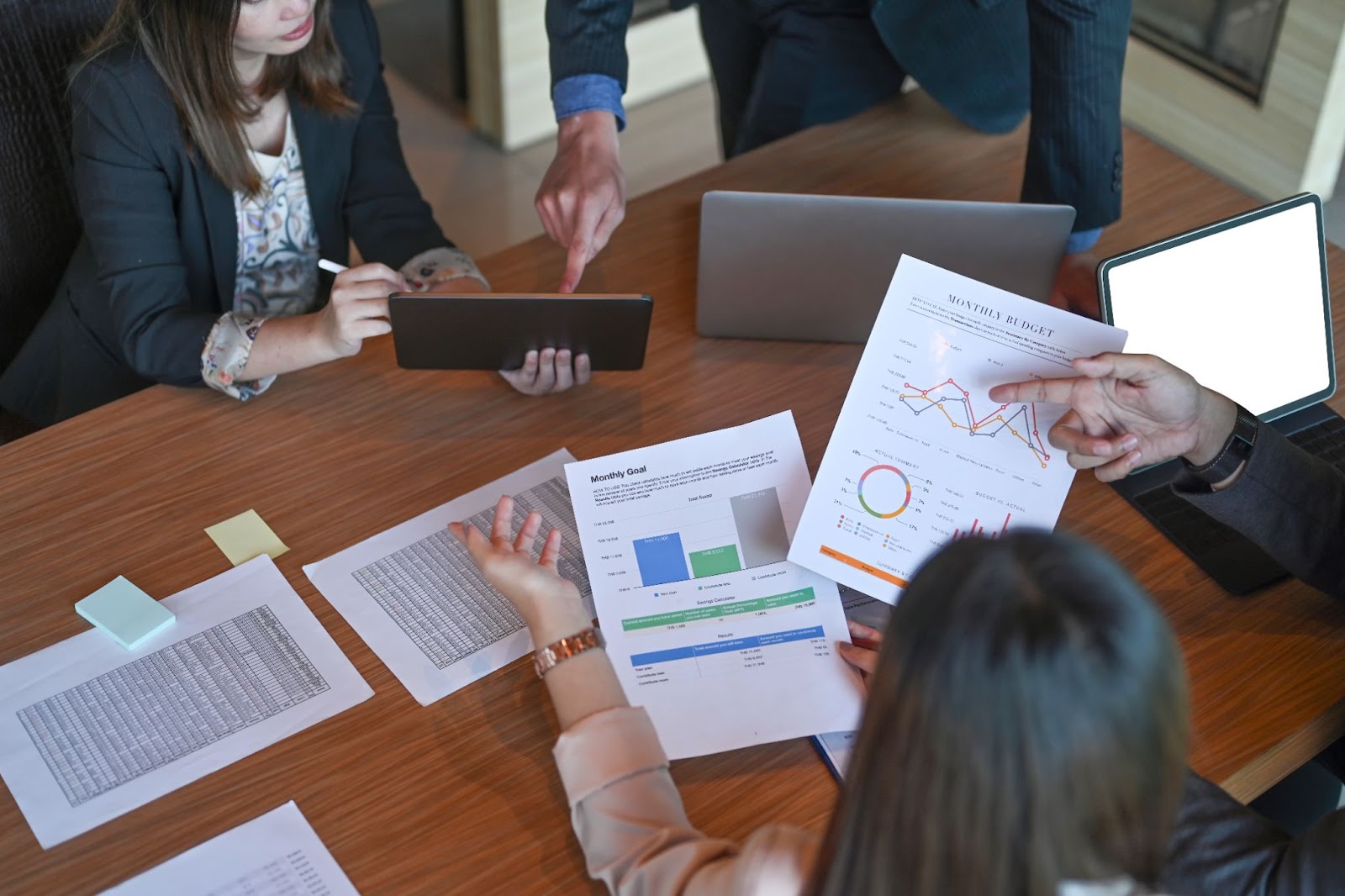 A cropped shot of businesspeople brainstorming and analyzing financial reports in a meeting room, with charts, a tablet, and laptops on the table.