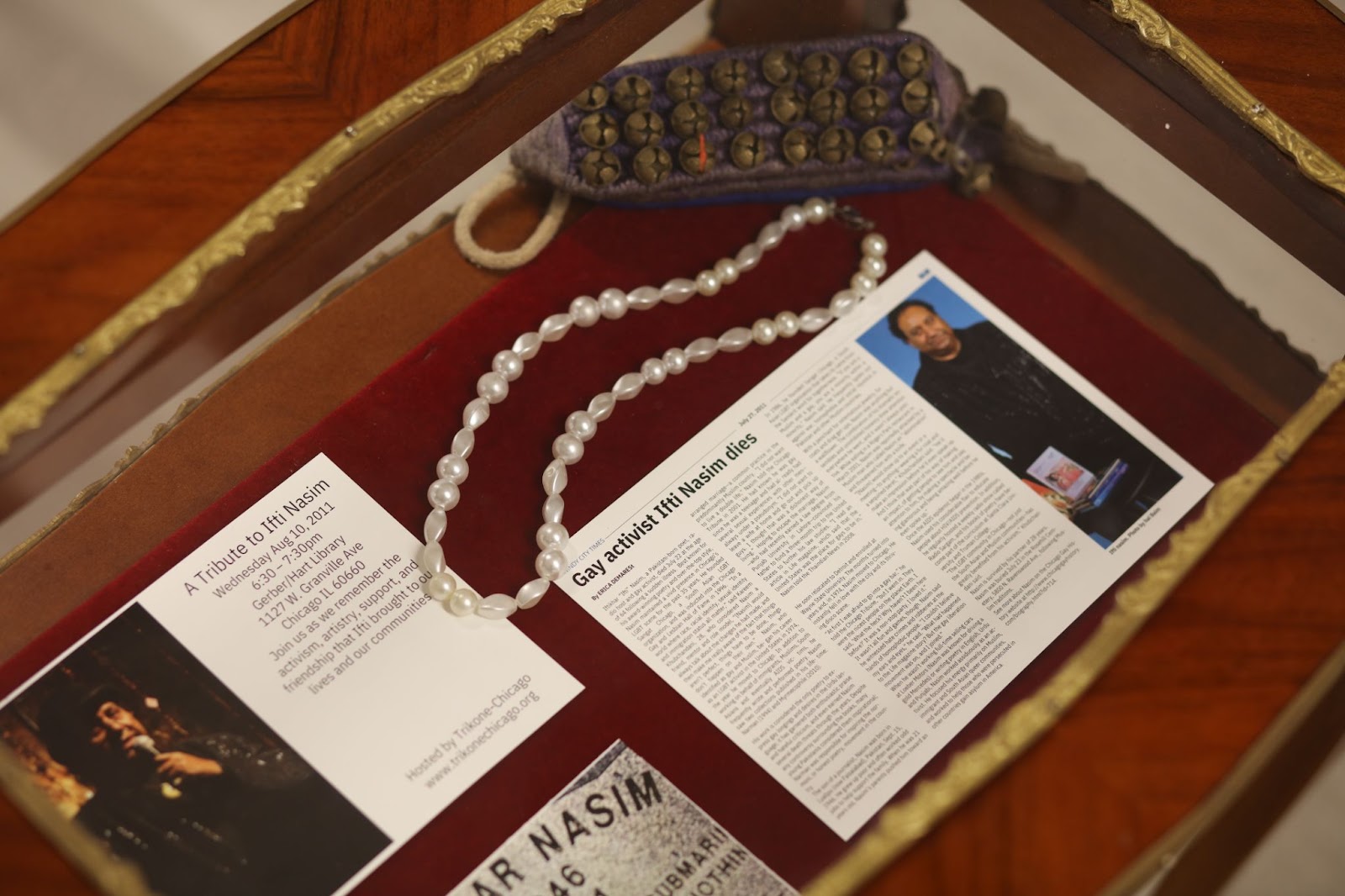 Image: Installation view, People Who Came Before Us. Encased in glass are a newspaper obituary of Ifti Nasim, an invitation to a tribute in his memory at the Gerber/Hart Library, a pearl necklace, and an item adorned with bells. Courtesy of South Asia Institute.