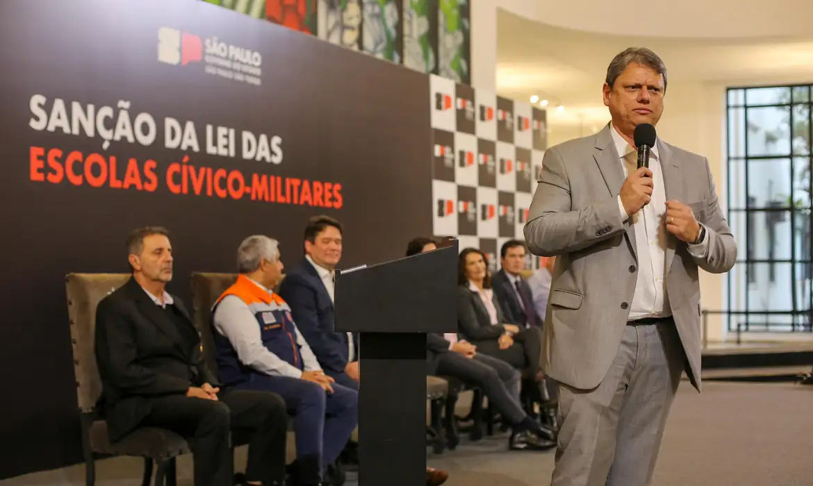 Governador Tarsicio de Freitas, de São Paulo, com um microfone na mão, em frente a um grupo de adultos sentados e um painel com a logo do governo e os dizeres "Sanção da lei das escolas cívico-militares"