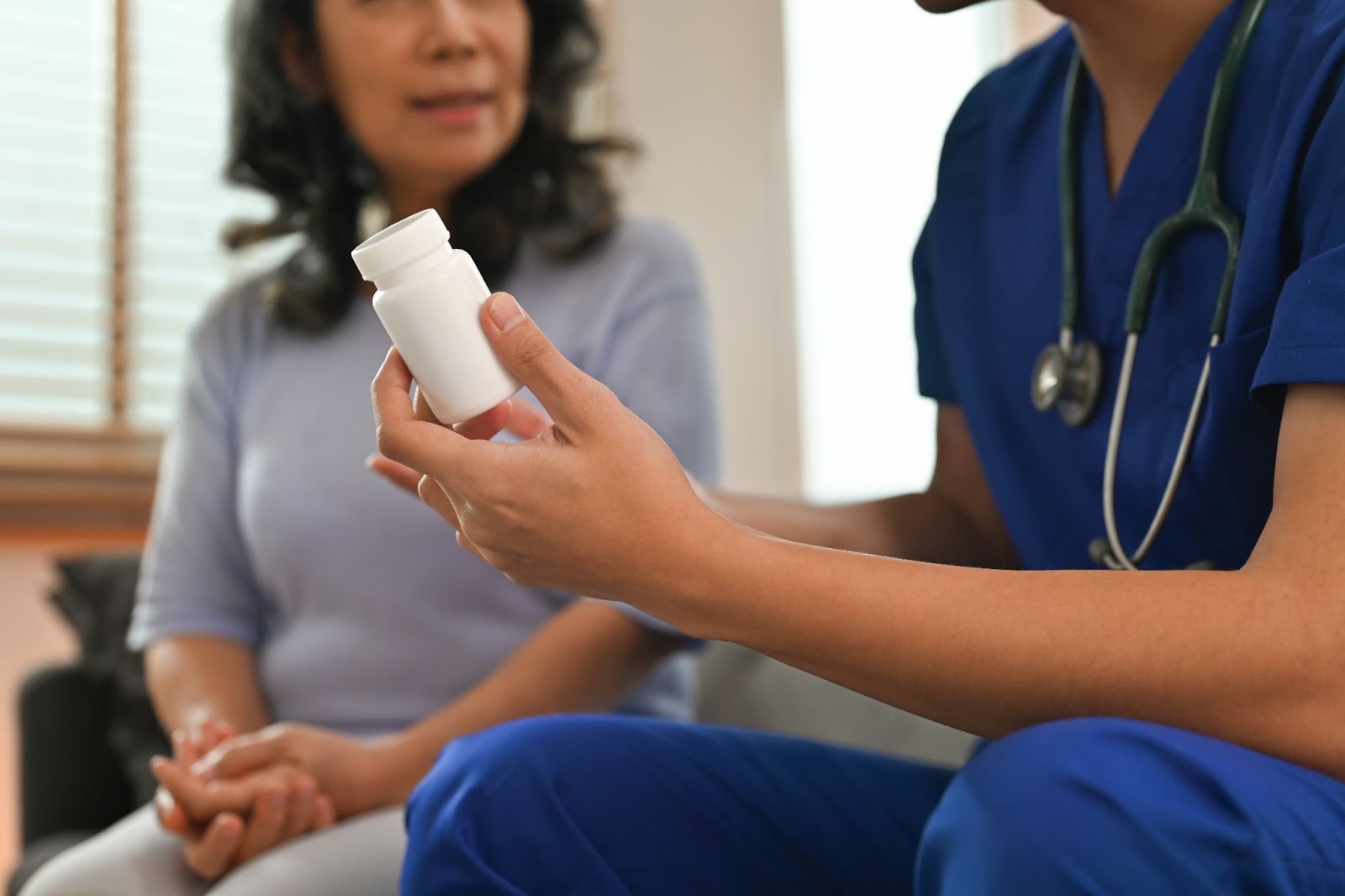 A cropped view of a female nurse’s hands holding a white bottle of pills next to a woman with dark hair blurred in the background.