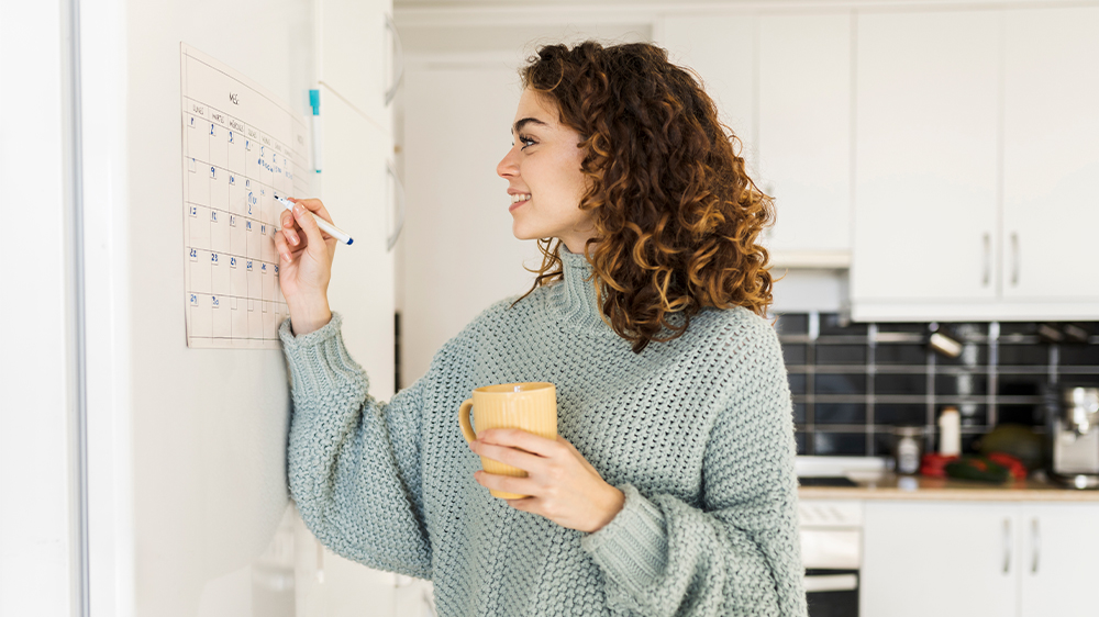 A woman with curly hair wearing a sweater holds a yellow mug and writes on a wall calendar in a modern kitchen with black tile backsplash and white cabinets. She is smiling softly.