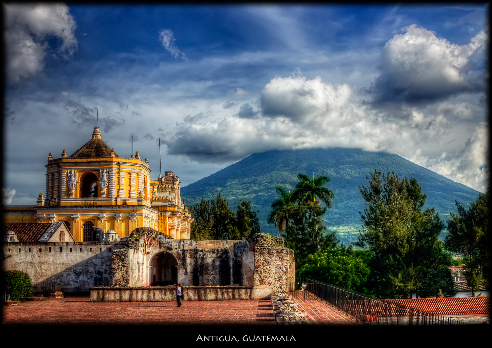 An attractive panorama of Agua, Guatemala, featuring lush vegetation, a tall volcano, and antique architecture beneath a foggy sky.