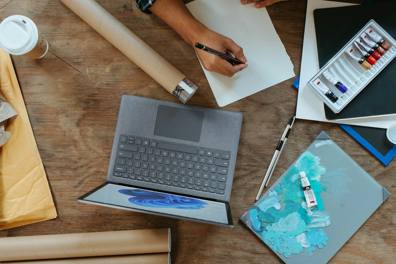 A man working on a table with laptop, paper, pencil and acrylic paint.