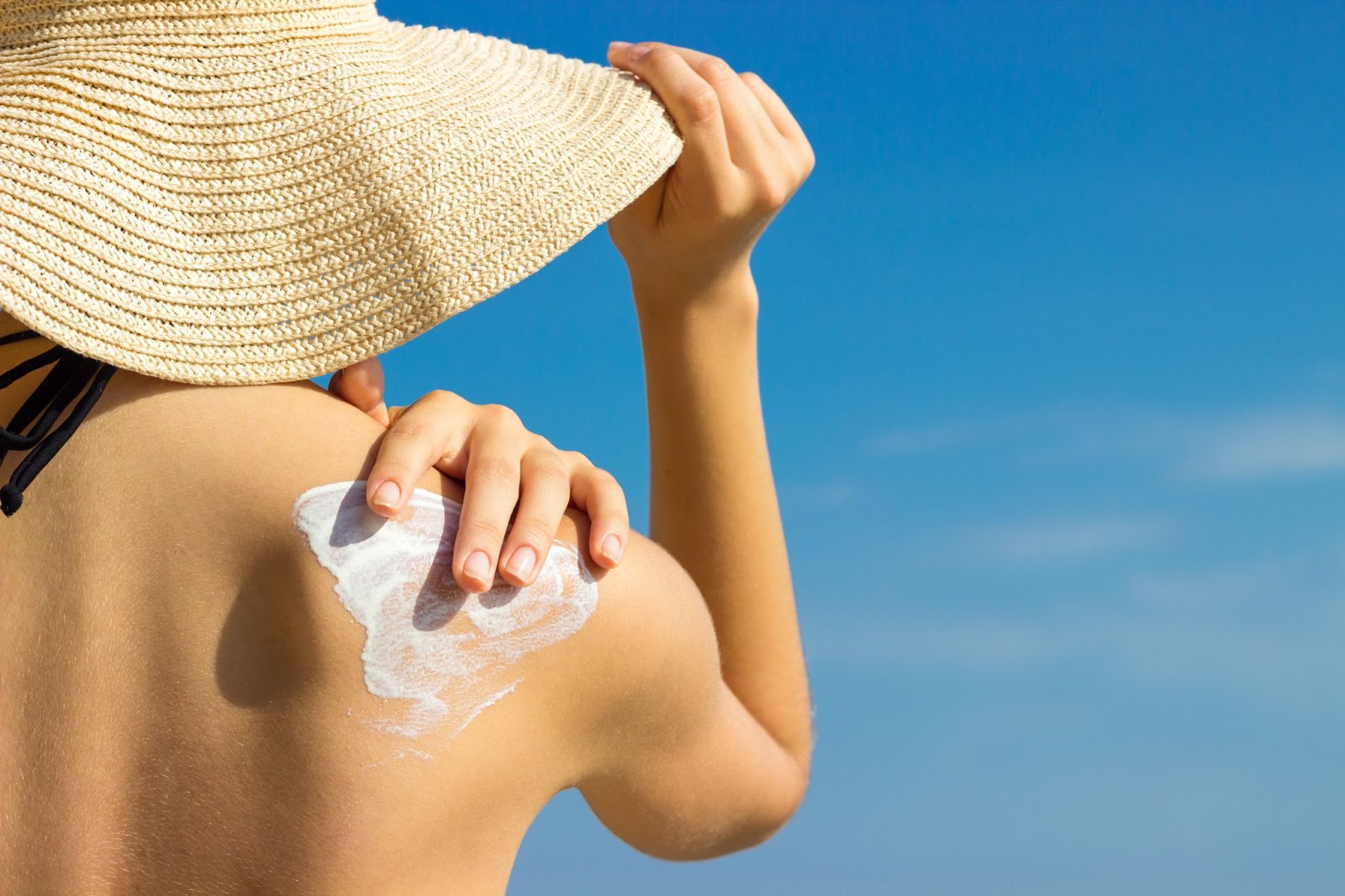 Woman applying sunscreen on her shoulder while wearing a wide-brimmed straw hat, emphasizing sun protection and skincare under a clear blue sky