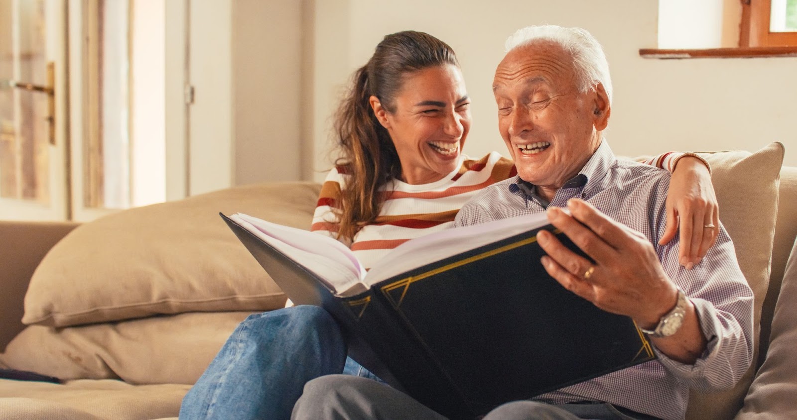 A senior man and his adult daughter laughing while looking at a photo album on their couch.