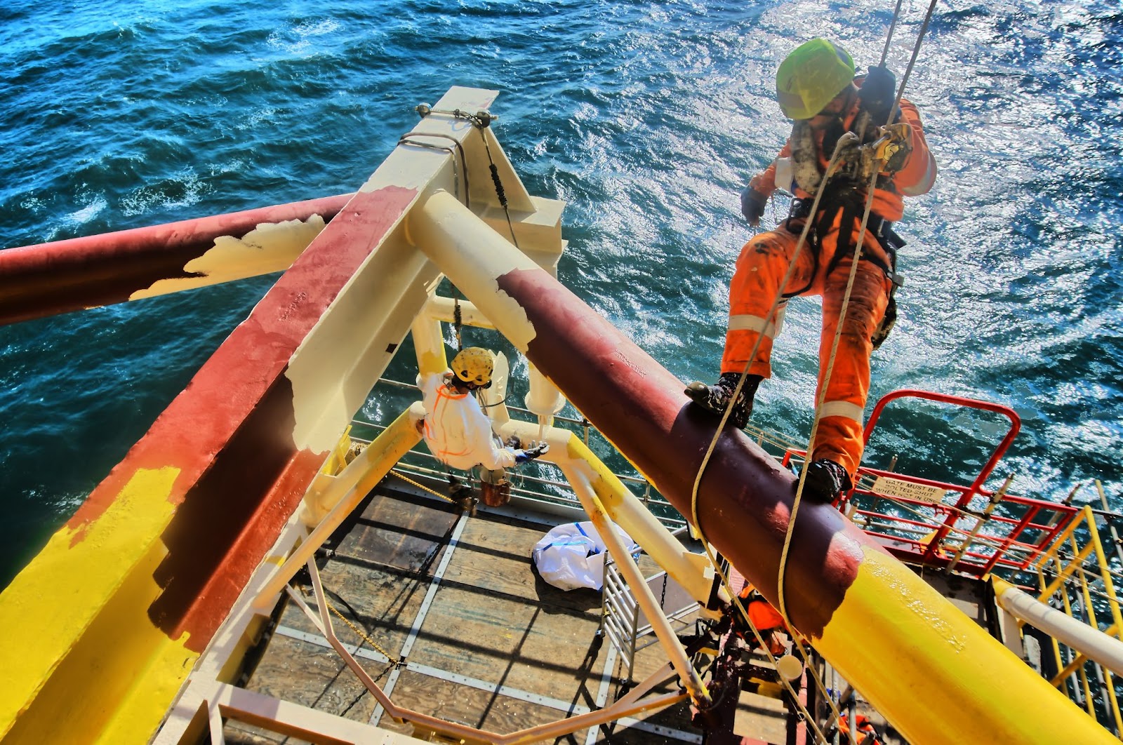 A worker in hi-vis overalls descends using industrial rope access onto a section of an offshore oil rig to paint it.