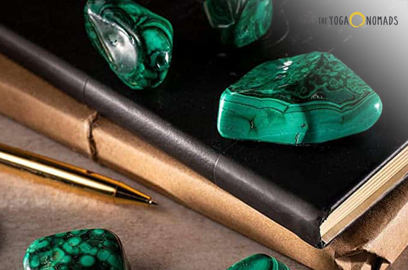 Several pieces of malachite, a green mineral with banded patterns, placed on and around a black book. A gold-colored pencil is also visible next to the book. The malachite’s rich green color and unique patterns make it visually striking.