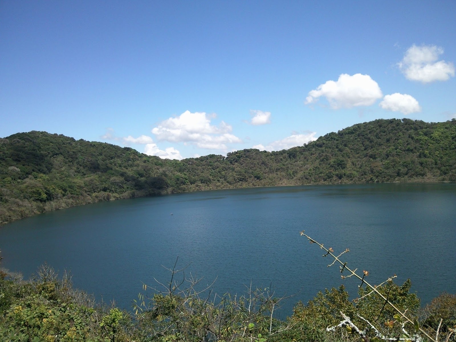 A lake encircled by Guatemala's Volcán Ipala. There is a clear sky, green forested hills, and clouds in the sky.