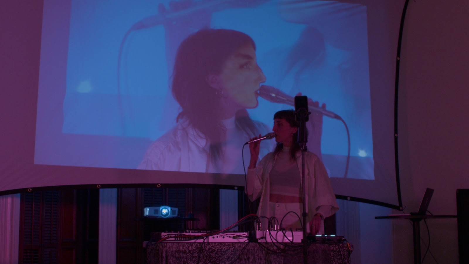 Image: Jessica Tucker performs “Possession” at PS1 Close House. Photo courtesy of the artist. A performer wearing all white stands behind a DJ booth. An iPhone is set up on a tripod in front of them. The performer is standing facing the left side of the image and singing into a microphone. A projector behind the performer displays an image of the performer facing the opposite direction while wearing an AR filter of the performer’s face. A larger projection facing the same direction as the performer is half-visible behind the first.