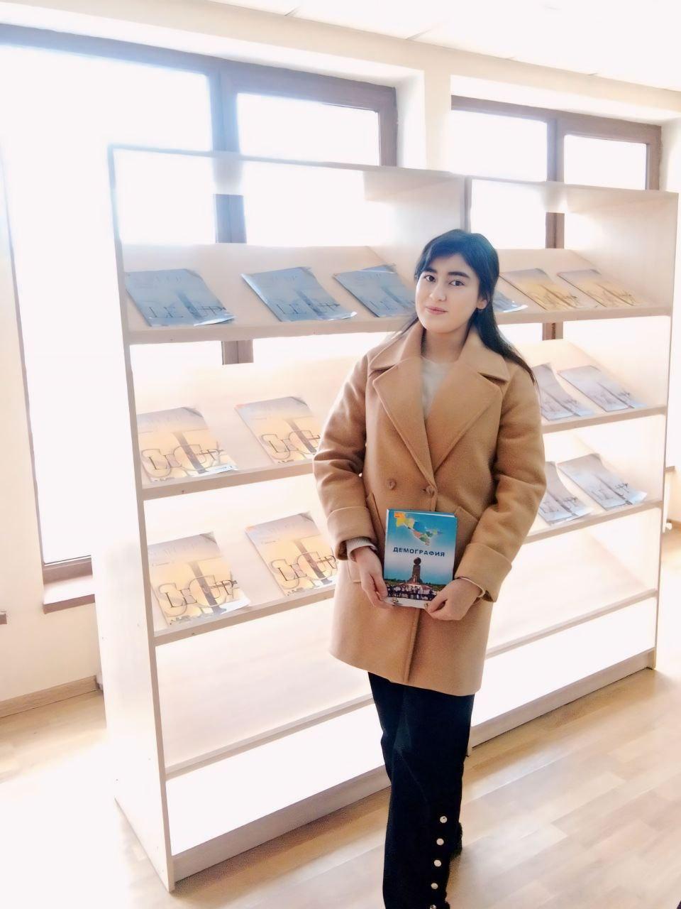 Young Central Asian woman holding a book and standing in front of a bookshelf. She's wearing a brown coat and black pants and has long straight dark hair. 