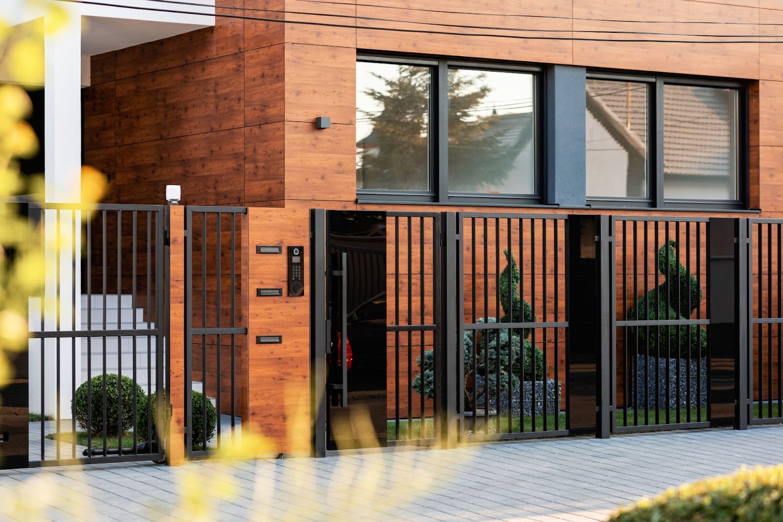Exterior view of a modern brown residential building featuring a sleek black steel gate adorned with two sculpted plants at the entrance.
