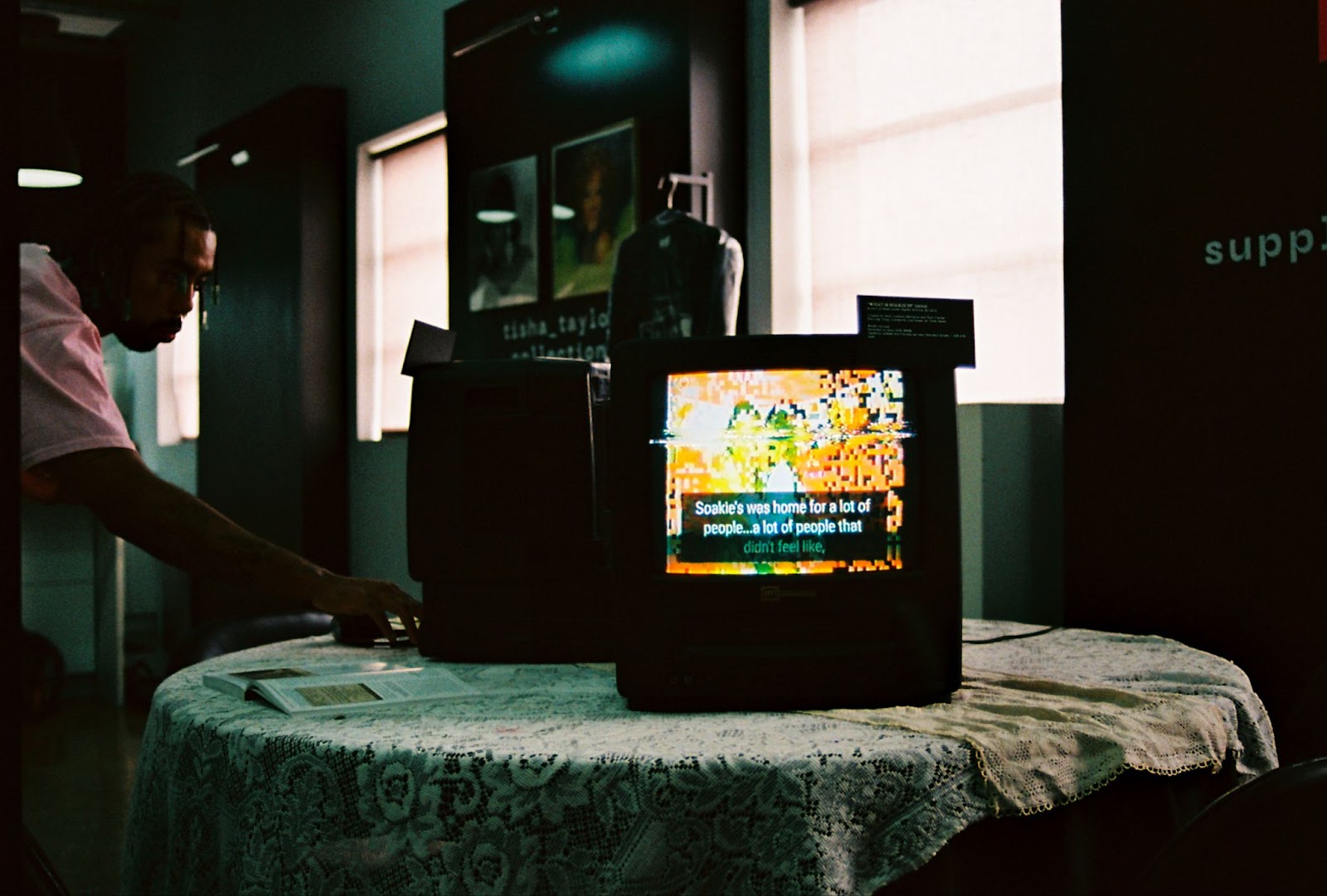 Image: Montalvo interacting with video installations featuring videos of Soakie’s Interviews displayed at Montalvo’s {B/qKC}: The Volume_2 Archive Launch Party. He reaches in to touch a TV. The table has a taffeta tablecloth and the room is dimly lit with a mint green tinge covering the room. Behind the table is a photo collection and a door. Photo by Jade Williams.