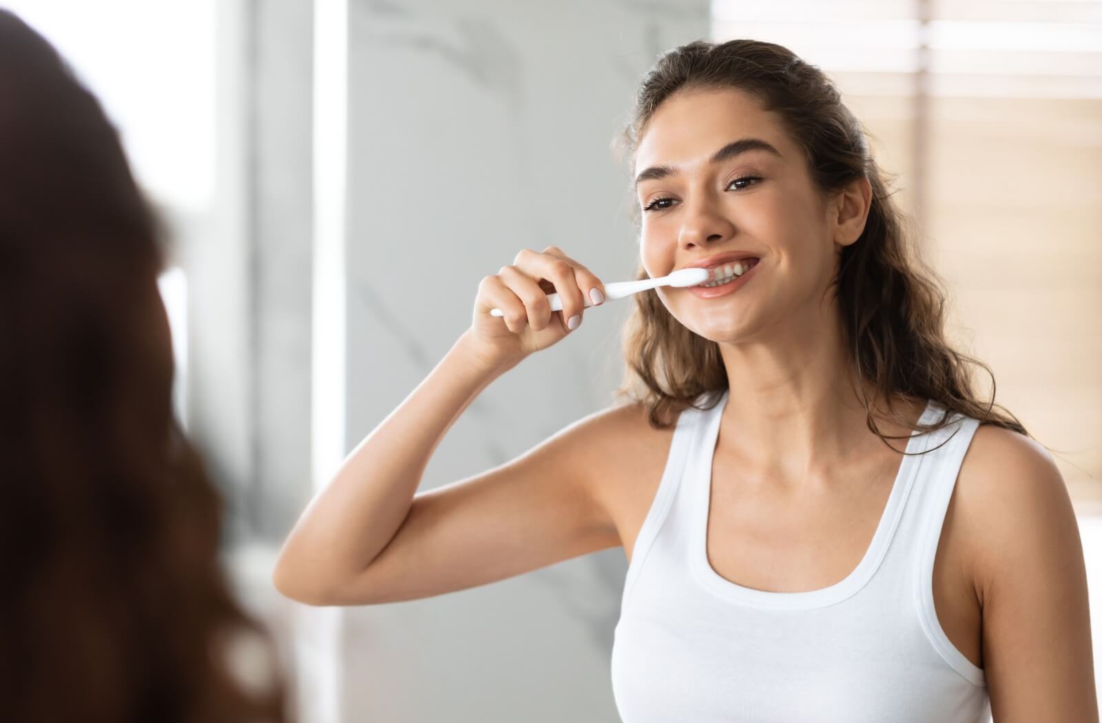 A woman in a white tank top brushing her teeth in front of a bathroom mirror.