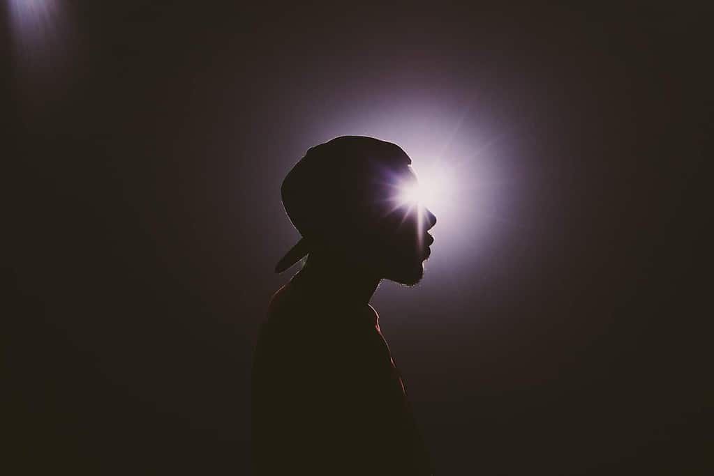 Ego death symptoms: The image depicts the silhouette of a person wearing a baseball cap, with bright light shining directly behind their head, creating a dramatic halo effect. The background is dark, emphasizing the stark contrast between the illuminated edges of the silhouette and the surrounding darkness.