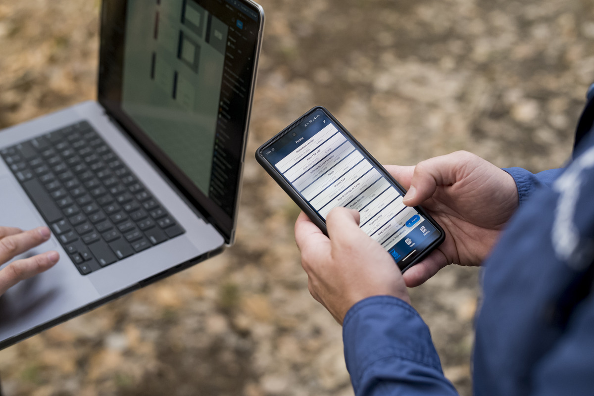 Person typing on a cell phone next to someone using a laptop. 