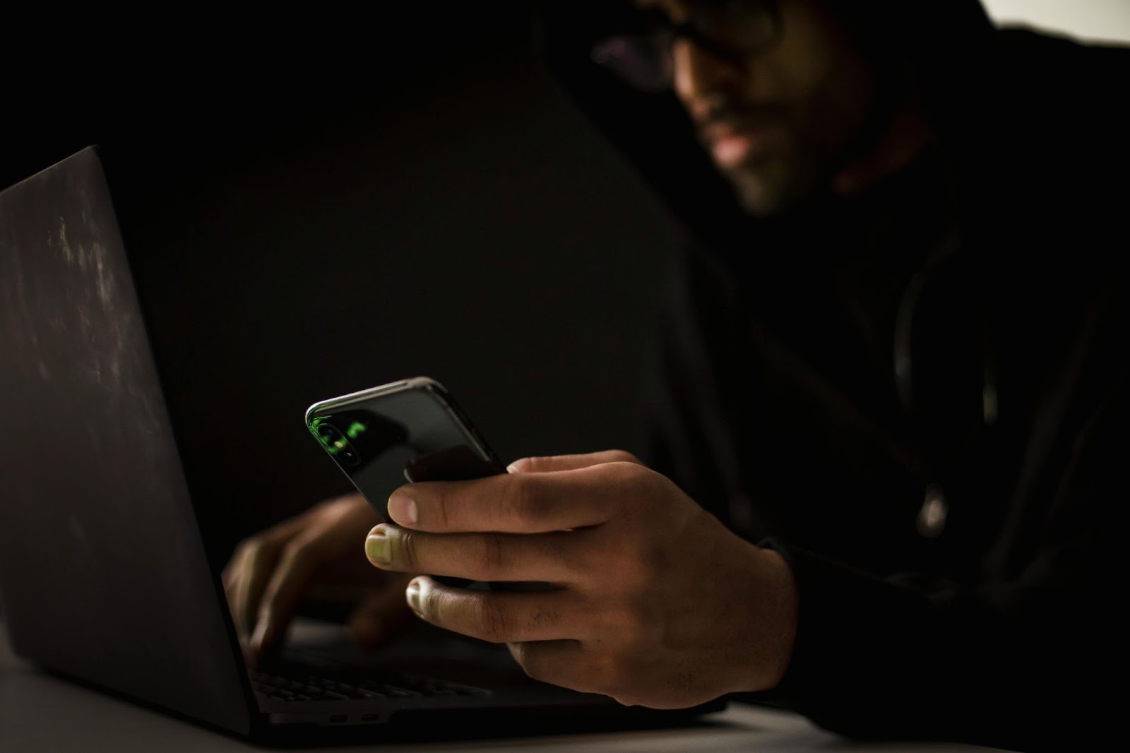 Man in a hoodie using a smartphone and laptop, symbolizing cyber blackmail and extortion activities