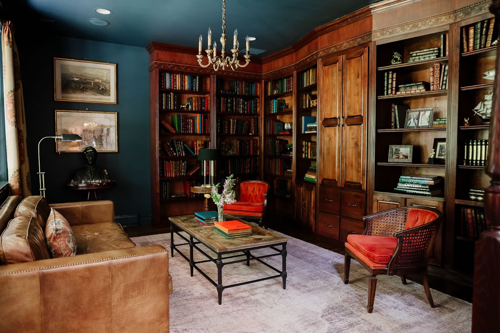 This library features built-in stained-wood shelving, a warm brown leather sofa and several other pieces of furniture and wall art on walls in dark, rich blue.