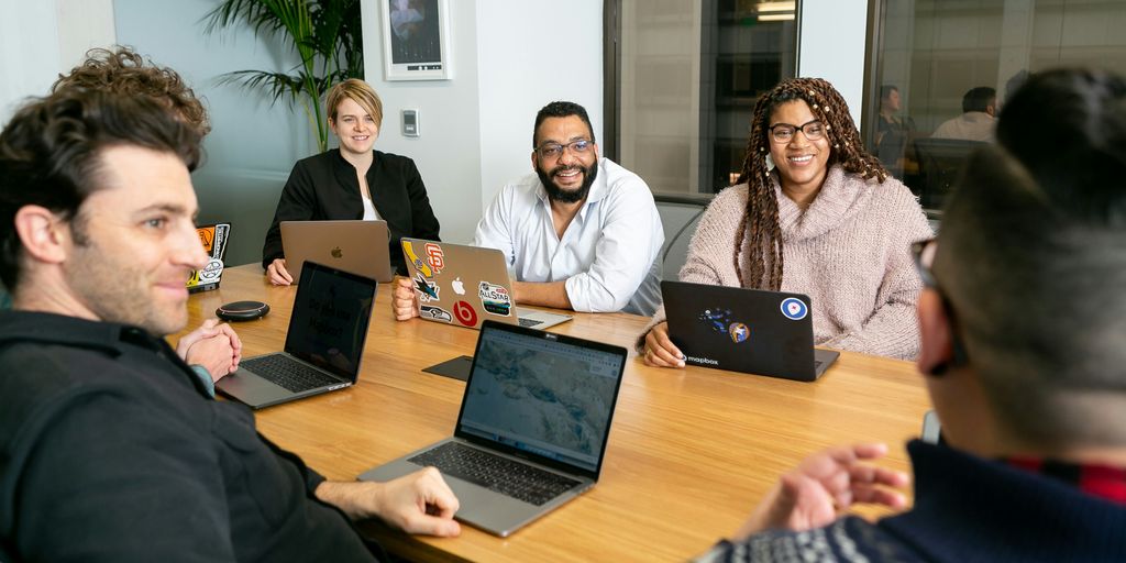 four people all on laptops, two men and two women, listen to person talking in a board meeting 