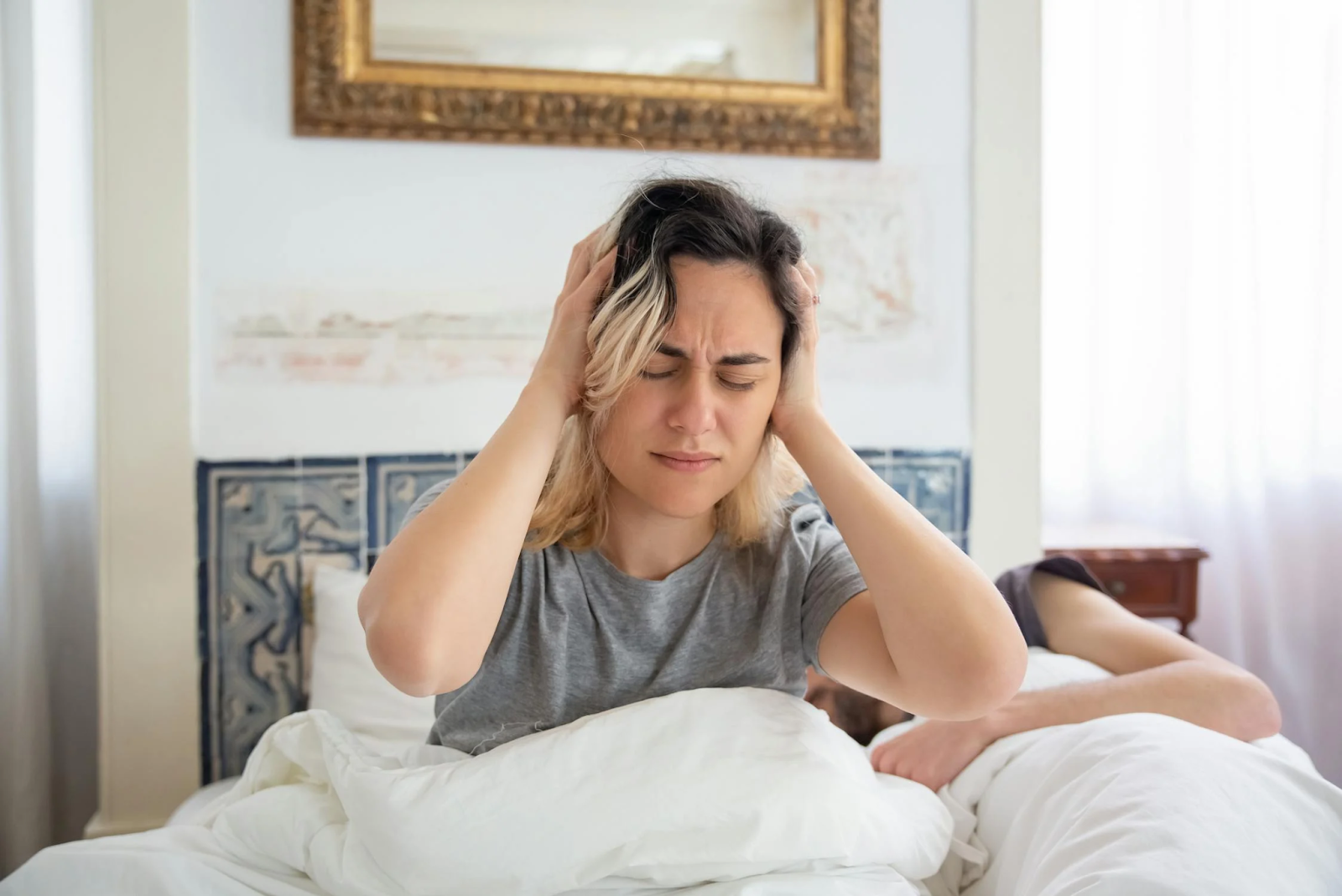 Woman in gray shirt holding her head while sitting on the bed. Headache can be effect of smoking on hangover.