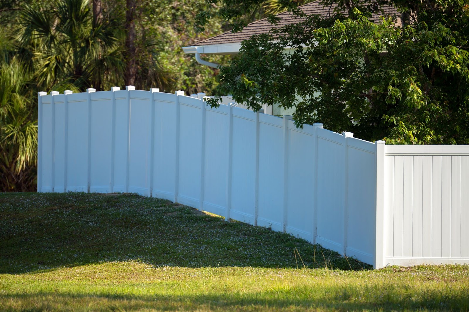 A white vinyl picket fence on a green lawn enclosing uneven property grounds.