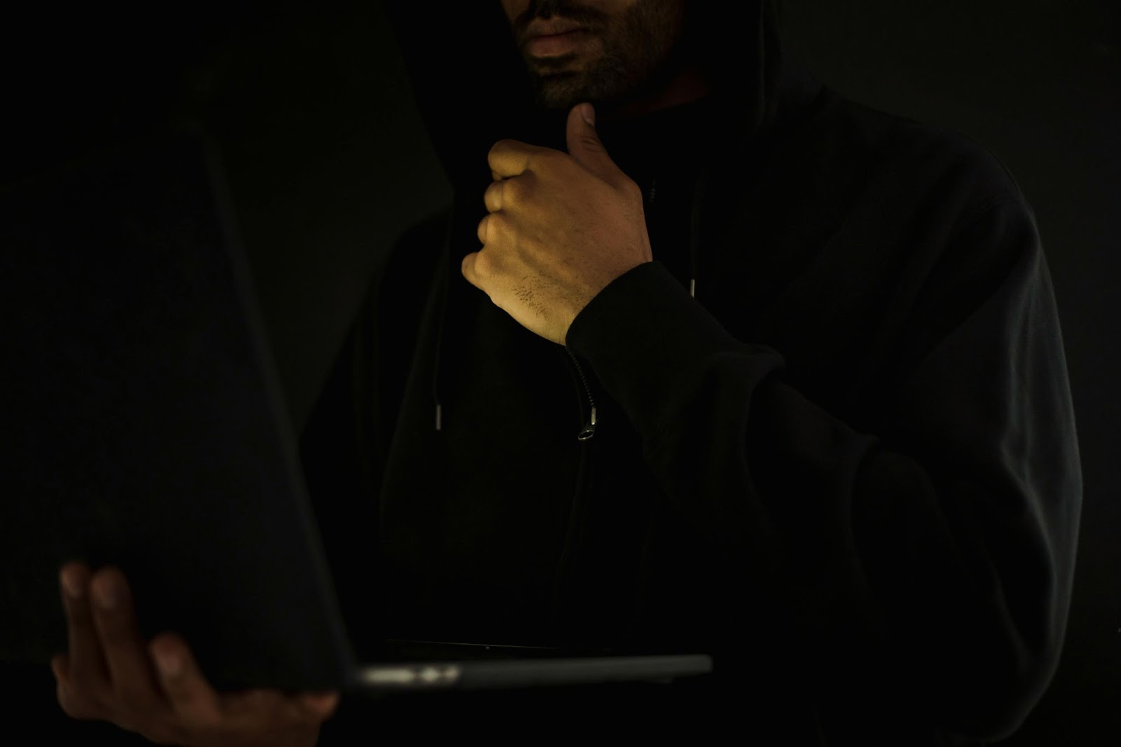 Man in a dark hoodie holding a laptop, evoking themes of secrecy in cyber blackmail and extortion