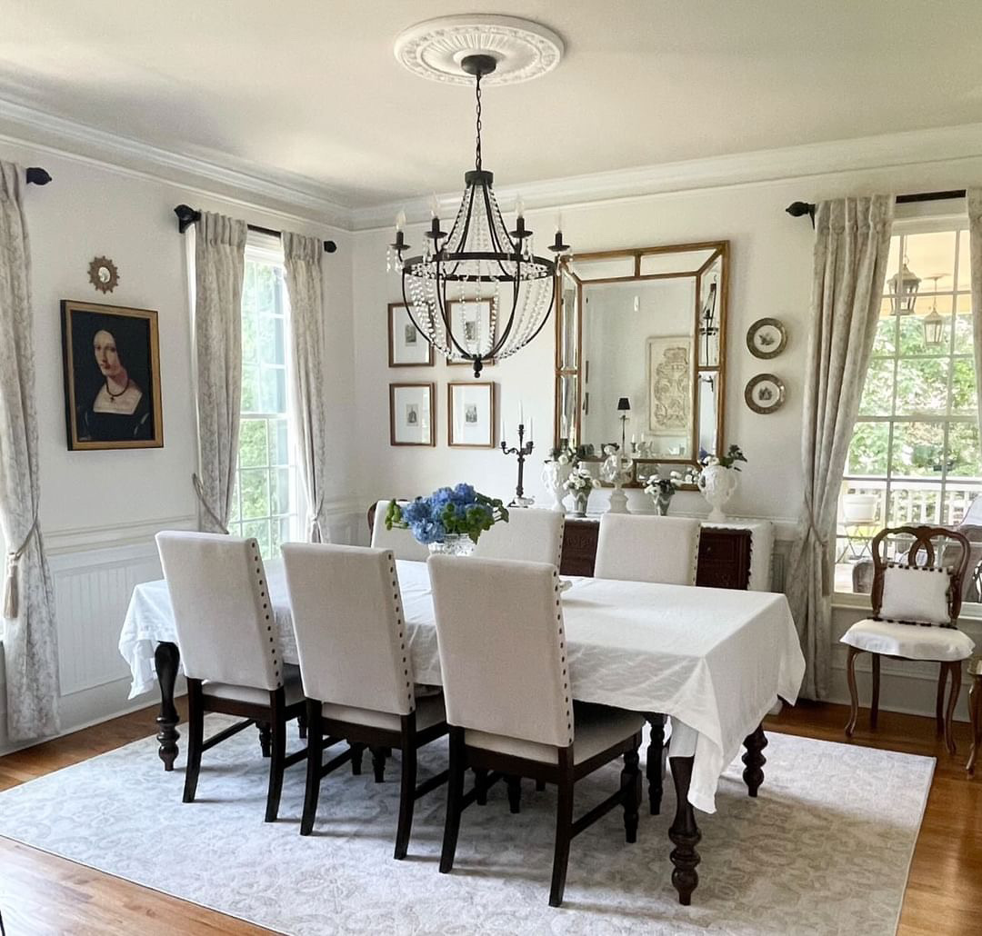 Nancy's dining room: Long table with white tablecloth & six dark wood chairs. Crystal chandelier & gold-framed mirror. Sideboard with candlesticks, vases & artwork. Hardwood floor, patterned rug & natural light.