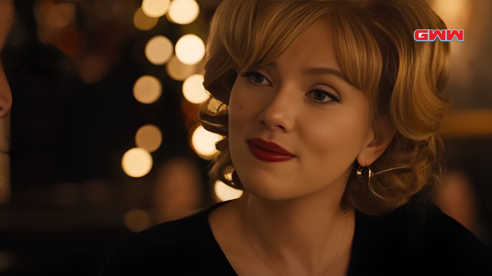 Kelly with curled blonde hair and red lipstick, smiling in a dimly lit setting