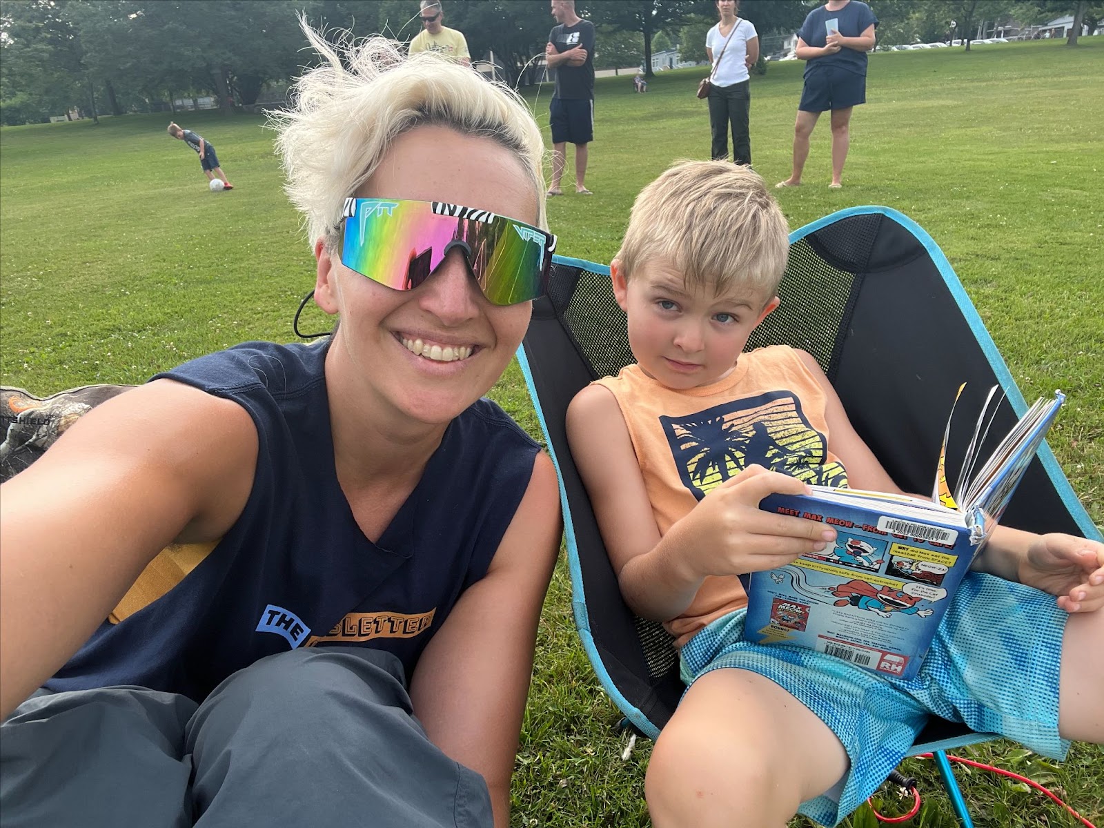 Tarzan at a soccer game with her son Mo. Tarzan wears a mirrored pair of Pit Viper sunglasses at a muscle-shirt that says "The Newsletter Conference." Her blond-haired son Mo sits in a Helinox camping chair with a book in his hand, raising his eyebrows at the camera.