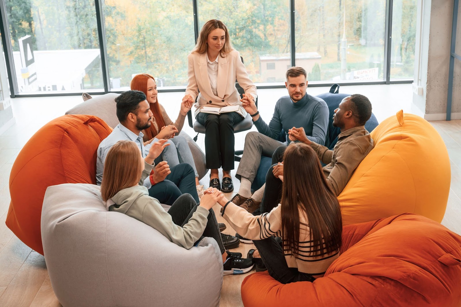 Group therapy with a therapist and several adults sitting on bean bag chairs while holding hands.